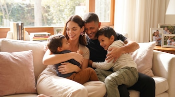 Una familia de cuatro, con Natalie Vértiz y Yaco Eskenazi, sentados en un sofá claro, abrazando a sus dos hijos en un ambiente hogareño y soleado.