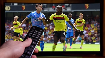Una mano sostiene un mando a distancia negro frente a una pantalla de televisión donde se transmite un partido de fútbol.