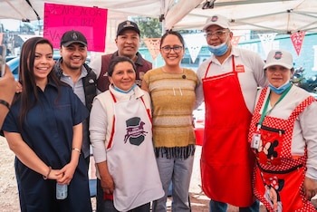 Grupo de siete personas sonrientes posando bajo una carpa blanca en un evento al aire libre. Llevan delantales y sombreros. Un letrero rosa dice 'BARBACOA LOS TRES'