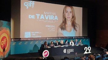 Pantalla grande con imagen de Marina de Tavira, texto 'Homenaje' y logotipos. Debajo, una mesa larga con varias personas sentadas y público