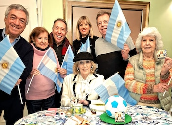 Siete personas adultas posan en un interior. Banderas de Argentina, una taza de té, una torta con forma de balón de fútbol, una tarjeta con imagen