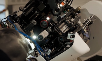 Primer plano de una máquina compleja con lentes, luces LED brillantes, cables azules y componentes electrónicos intrincados. Tonos oscuros y reflejos de luz