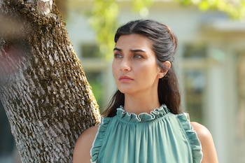 Primer plano de Tuba Büyüküstün con cabello oscuro recogido, luciendo una blusa verde con volantes blancos, apoyada en el tronco rugoso de un árbol