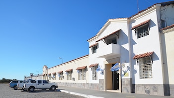 Fachada de edificio institucional blanco con ventanas enrejadas y toldos de tejas. Varias camionetas estacionadas. Cielo azul despejado