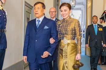 Un hombre en traje azul y una mujer en un vestido de noche dorado intrincado, con un cinturón enjoyado, caminan por un pasillo, rodeados de personal