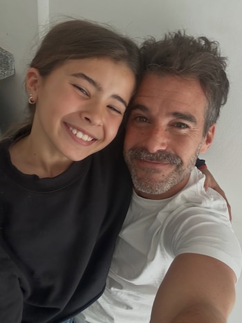 Hombre y niña posan abrazados sonriendo para una selfie, ambos de medio cuerpo. El hombre tiene barba y viste camiseta blanca. La niña lleva jersey negro
