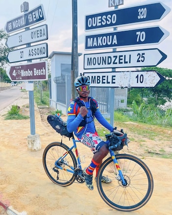 Un hombre con casco y gafas deportivas sobre una bicicleta azul, ubicado frente a múltiples carteles viales que señalan distancias a ciudades