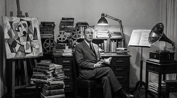 Fotografía en blanco y negro de un hombre sentado con un libro, junto a pilas de libros, un cuadro cubista, bobinas de cine y un gramófono.
