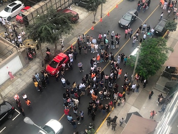 Vista aérea de una calle con un gran grupo de personas en la vía y aceras, varios coches y motocicletas aparcados y detenidos, y edificios