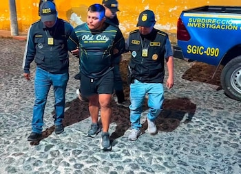 Dos agentes de policía con uniforme y gorras DEIC escoltan a un hombre en camiseta y shorts. El grupo camina sobre adoquines junto a un vehículo policial azul
