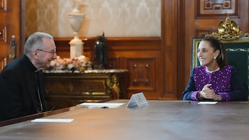 Un hombre con traje negro y clergyman sentado frente a una mujer con saco morado, ambos sonríen en una mesa de madera. Fondo con paneles de madera y muebles
