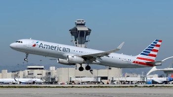 Un avión blanco de American Airlines con cola de rayas rojas y azules despega. La torre de control y otros aviones se ven en el fondo