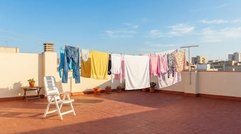 Ropa tendida en cuerdas para secar en una terraza de azotea con baldosas rojas. Se ven vaqueros, toallas y camisetas, una silla blanca y plantas.