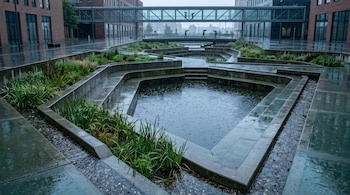 Plaza de hormigón con cuencas escalonadas de agua de lluvia, vegetación perimetral y peatones con paraguas sobre un puente al fondo.
