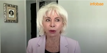 Mujer de cabello rubio corto y platino, con blazer lila, mirando hacia la derecha con boca abierta. Fondo de pared clara y cuadro decorativo