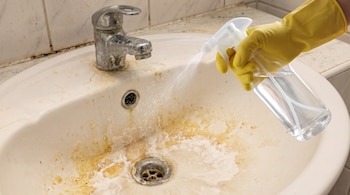 Una mano con guante amarillo sostiene una botella pulverizadora transparente que rocía líquido sobre un lavabo blanco muy sucio con grifo oxidado y manchas de cal y óxido.
