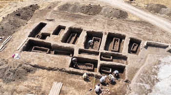 Vista aérea de un sitio arqueológico con varios arqueólogos excavando tumbas de tierra que contienen sarcófagos y esqueletos. Se ven montículos de tierra y un camino.