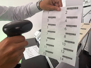Una mano sostiene un escáner de códigos de barras. La otra mano sujeta una hoja impresa con catorce códigos de barras numerados. Se observa una impresora