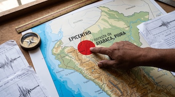 Una mano señala un círculo rojo sobre un mapa de Perú, rodeado por una brújula, una regla de madera y hojas con gráficos de ondas sísmicas.