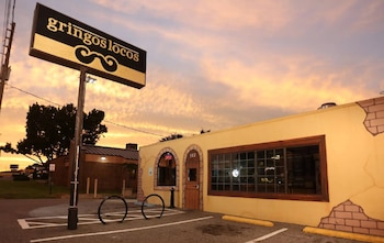 Letrero de Gringos Locos con logo de bigote. Edificio amarillo con puerta de madera, ventana, rótulo luminoso OPEN y número 517. Aparcabicicletas en estacionamiento. Cielo atardecer