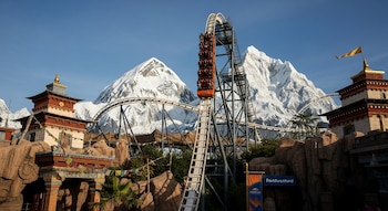 Un tren naranja lleno de pasajeros asciende la empinada cuesta de una montaña rusa, flanqueado por estructuras temáticas himalayas y montañas nevadas falsas.