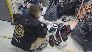Persona con chaqueta negra de UTOJ y Min Seg Caba, examinando zapatillas en el suelo junto a bolsas de basura negras en un local