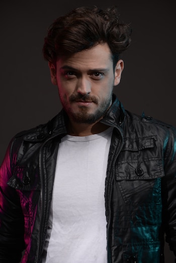Hombre con cabello oscuro y barba, chaqueta negra y camiseta blanca. Iluminación azul y magenta en rostro y chaqueta. Fondo oscuro