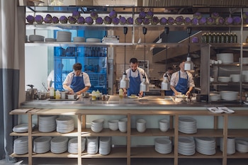 Vista frontal de tres chefs en chaquetas blancas y delantales azules, preparando alimentos en una cocina de restaurante moderna, con estantes de platos y alcachofas decorativas