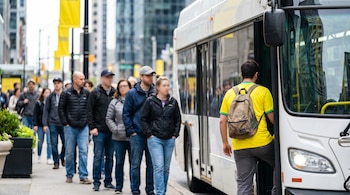Hombre con camiseta amarilla y mochila subiendo a un bus blanco. Detrás, una fila de personas en una calle urbana con edificios desenfocados.