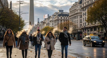 Cuatro personas abrigadas caminan por una vereda con hojas secas, exhalando vapor en el aire frío, con el Obelisco de Buenos Aires y edificios a la derecha, y un taxi en la calle.