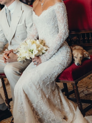 Charles Leclerc y Alexandra Saint Mleux se toman de las manos en su boda. Ella lleva un vestido de encaje blanco y un ramo de lirios. Un perro marrón claro duerme junto a ellos