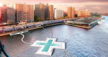 Vista aérea de una piscina flotante con forma de cruz en el East River de Nueva York, conectada a la orilla con edificios y una autopista