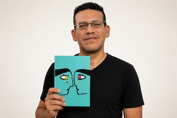 Un hombre con gafas y camiseta negra sostiene un libro de color turquesa con la ilustración de dos rostros enfrentados.