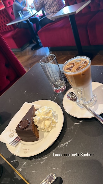 Rebanada de torta Sacher con sello de chocolate y crema batida, un café helado y un vaso sobre mesa oscura. Al fondo, sillones rojos de una cafetería