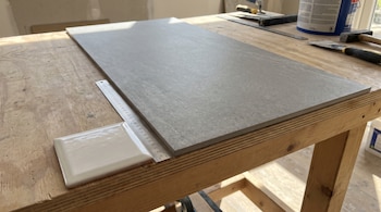 Un azulejo blanco pequeño, una regla metálica y una placa cerámica gris grande sobre una mesa de madera con herramientas de construcción.