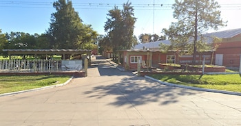 Un camino asfaltado conduce hacia los edificios de paredes rojas de una fábrica, con árboles y un cartel que dice Algodonera Avellaneda