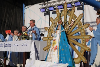 Una escultura de la Virgen de Luján en primer plano frente a sacerdotes con vestimentas celestes y blancas en un altar