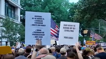 Multitud de personas en una manifestación al aire libre, sosteniendo pancartas con texto y banderas de Estados Unidos e Israel