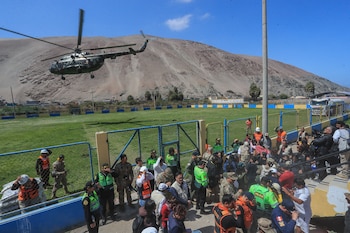 Un helicóptero sobrevuela un campo de fútbol con cerco metálico rodeado de policías, rescatistas y pobladores al pie de un cerro