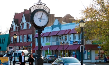 Una calle urbana en Lakewood con un reloj público en el centro, edificios de ladrillo y fachadas claras, personas caminando, coches y árboles con hojas de otoño