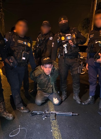 Un hombre esposado y arrodillado frente a varios agentes de policía en una calle oscura, con un fusil de asalto negro en el suelo