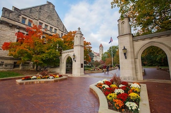 Vista de las Sample Gates de la Universidad de Indiana con edificios de piedra, árboles otoñales y caminos de ladrillo. Parterres con flores de colores