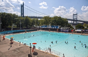 Gran piscina azul al aire libre con bañistas y socorristas con sombrillas naranjas. El Puente Robert F. Kennedy y árboles verdes de fondo bajo un cielo parcialmente nublado