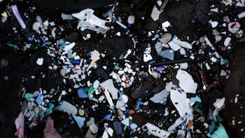 Primer plano de una superficie oscura cubierta con innumerables piezas de plástico roto en tonos blanco, azul, verde, rosa y marrón, mezcladas con pequeñas rocas