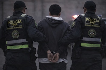 Vista trasera de un joven esposado, con buzo oscuro, escoltado por dos agentes de la PNP con chalecos reflectivos y uniformes policiales.