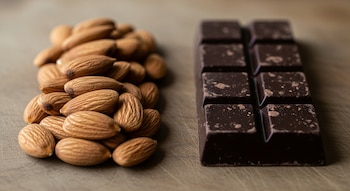 Una pila de almendras sin cáscara se encuentra a la izquierda, junto a una barra de chocolate negro dividida en cuadrados a la derecha, sobre una tabla de madera.