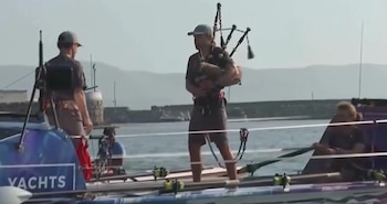 Un hombre tocando la gaita en un bote, acompañado por dos personas más. El bote está en el agua, con un paisaje montañoso y el cielo claro de fondo