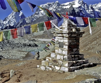 Memorial de piedra gris adornado con banderas de oración de colores. Al fondo, un lago azul verdoso y cumbres rocosas nevadas bajo un cielo azul