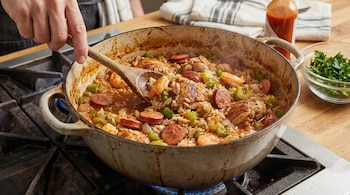 Mano revolviendo un guiso de jambalaya en una olla grande sobre una estufa de gas, con arroz, trozos de pollo, salchichas, camarones y pimientos verdes.
