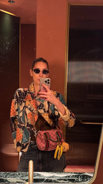 Una mujer con gafas de sol y chaqueta estampada se toma una foto frente a un espejo con un teléfono móvil y lleva un bolso rosado cruzado